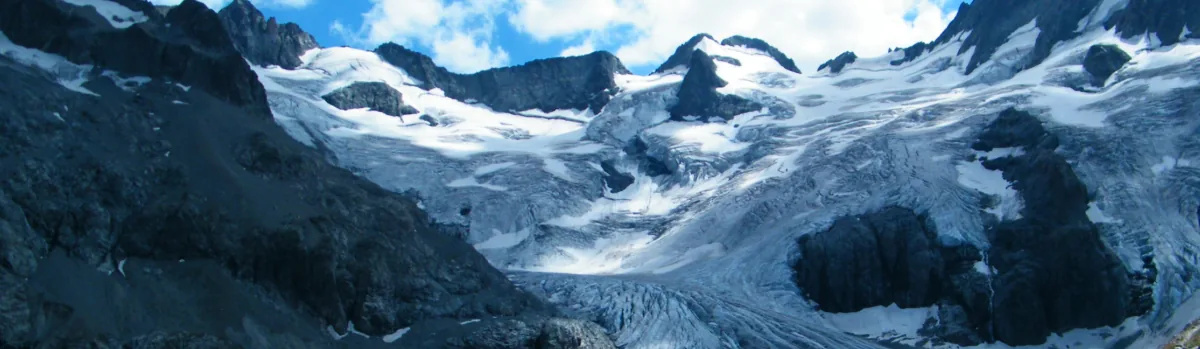 Detail of a photo: Glacier with curved grey stripes flowing down a valley from snowy, rocky peaks under a blue sky with white clouds, beside a gravel path and a wooden fenced viewpoint.