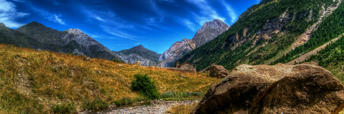Detail of a photo: Large angular boulder beside a shallow pebbly stream in a grassy mountain valley, below steep forested slopes and rocky peaks under a dramatic blue sky, in heavily processed colours.