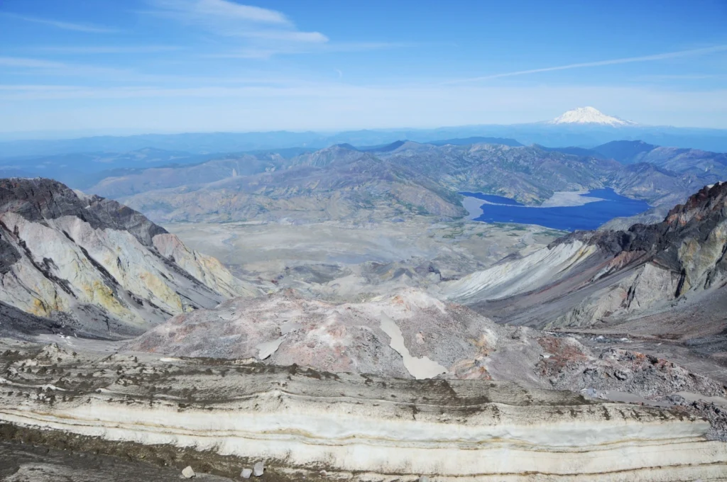 The view down over Mount St. Helens crater from the summit, in the centre the lava dome has grown in the collapse scar from the 1980 eruption. The collapse devastated the proximal land and vegetation, dead trees still float like matchsticks in the calm waters of Spirit Lake and the event left the inner workings of the volcano open to scrutiny. In the background, the glacier-capped Mount Rainier lies dormant. (Credit: Jackie Kendrick)