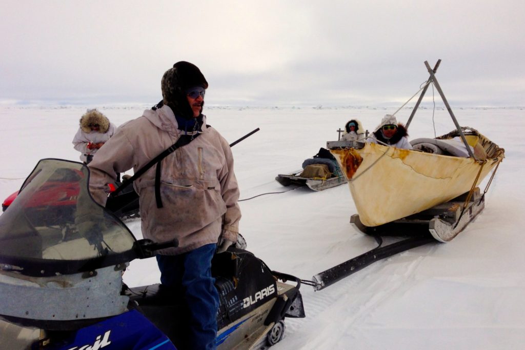 Man on snow machine looks bake to the canoe he is towing across ice.