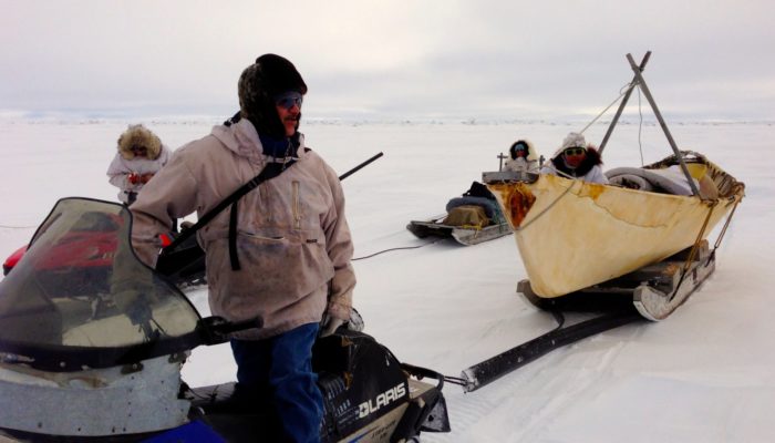 Man on snow machine looks bake to the canoe he is towing across ice.