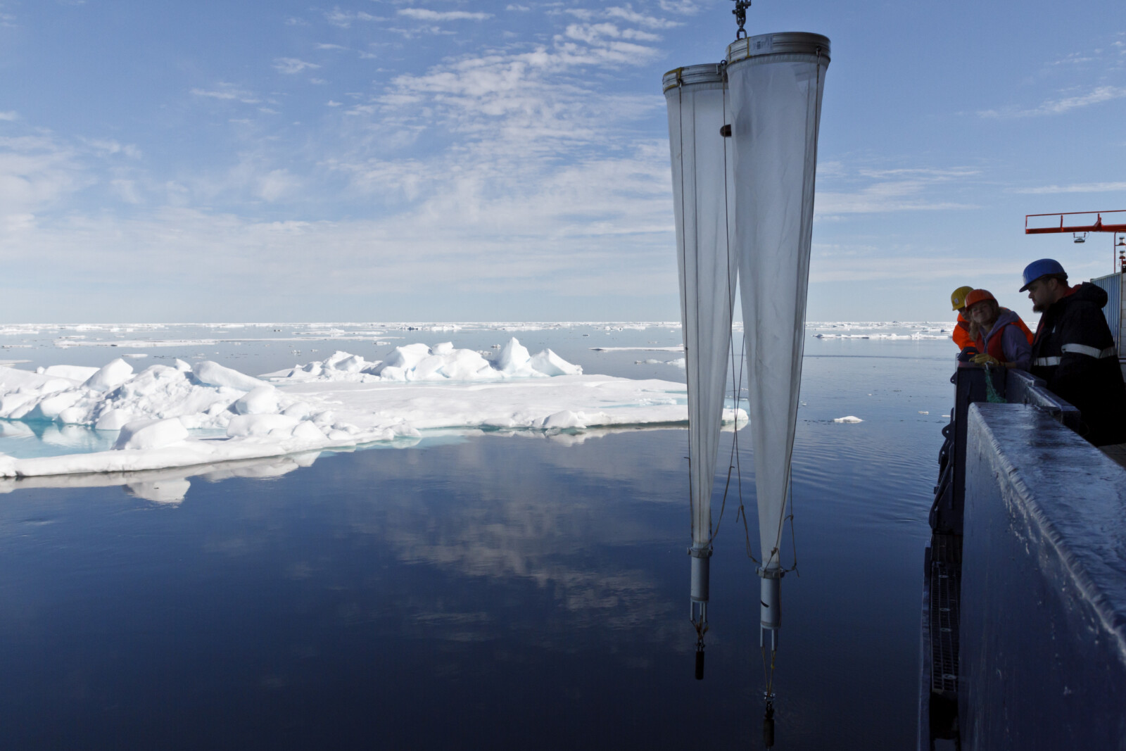 In front of blue skies and some floating sea ice, long cylindrical nets are hauled back to a research boat. On the right, three people with helmets wait to get the nets in.