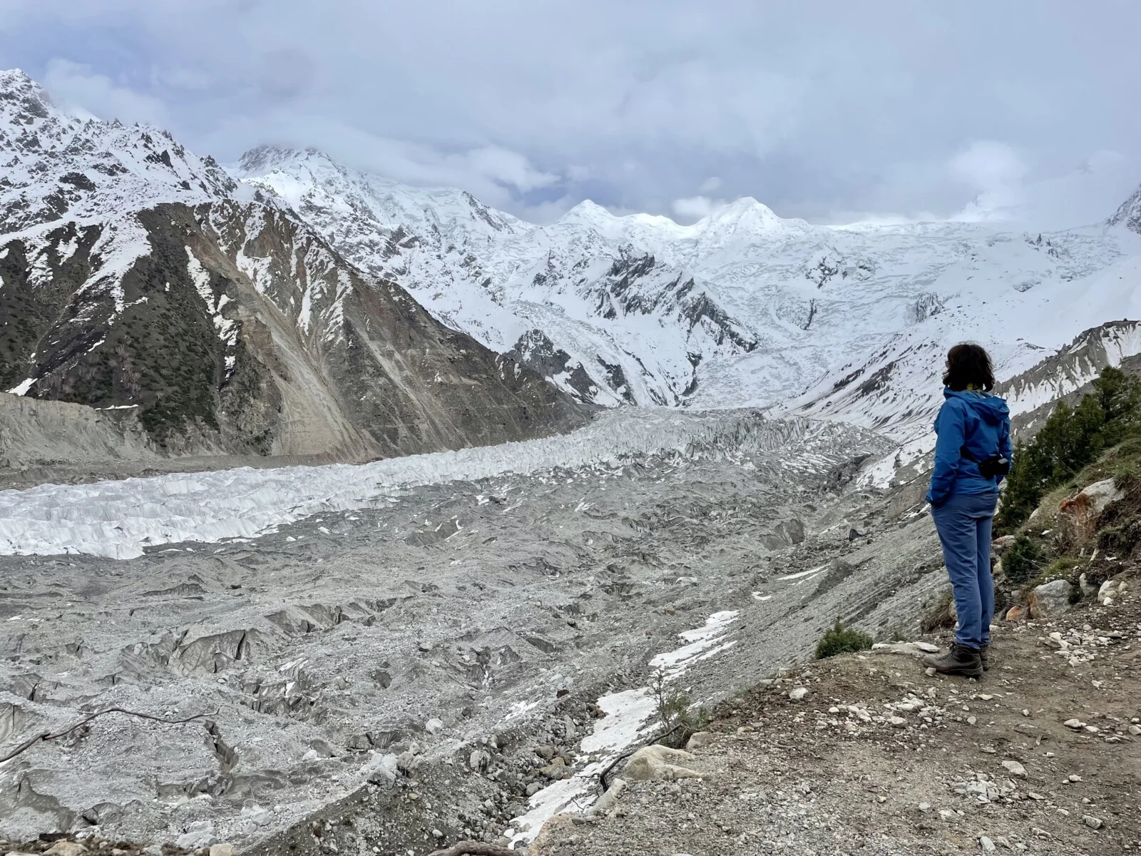 Alex stood next to a glacier in the Karakoram region. The glacier is debris coloured and in the background there are mountains covered in snow. 