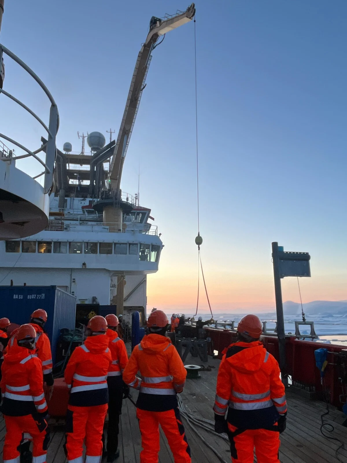 Glaciologists stood on the deck of the RSS Sir David Attenborough on a cruise to the Arctic with the sun setting in the background and glaciers in the ocean. 