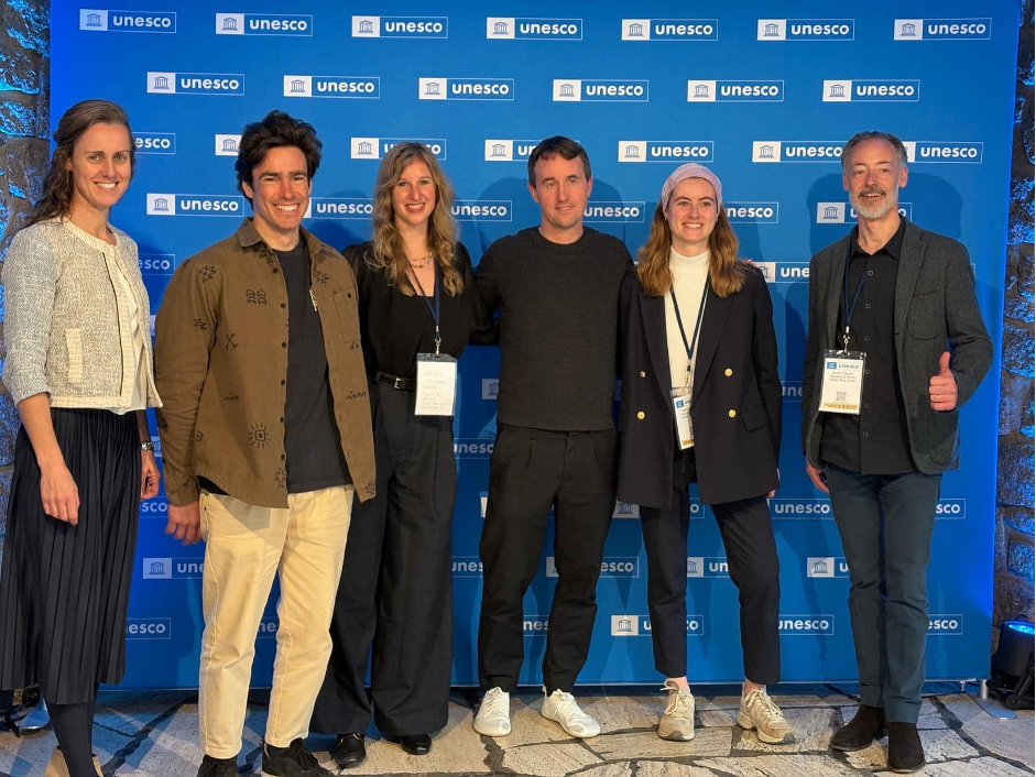six individuals stood in font of a blue wall with the UNESCO logo on it. 