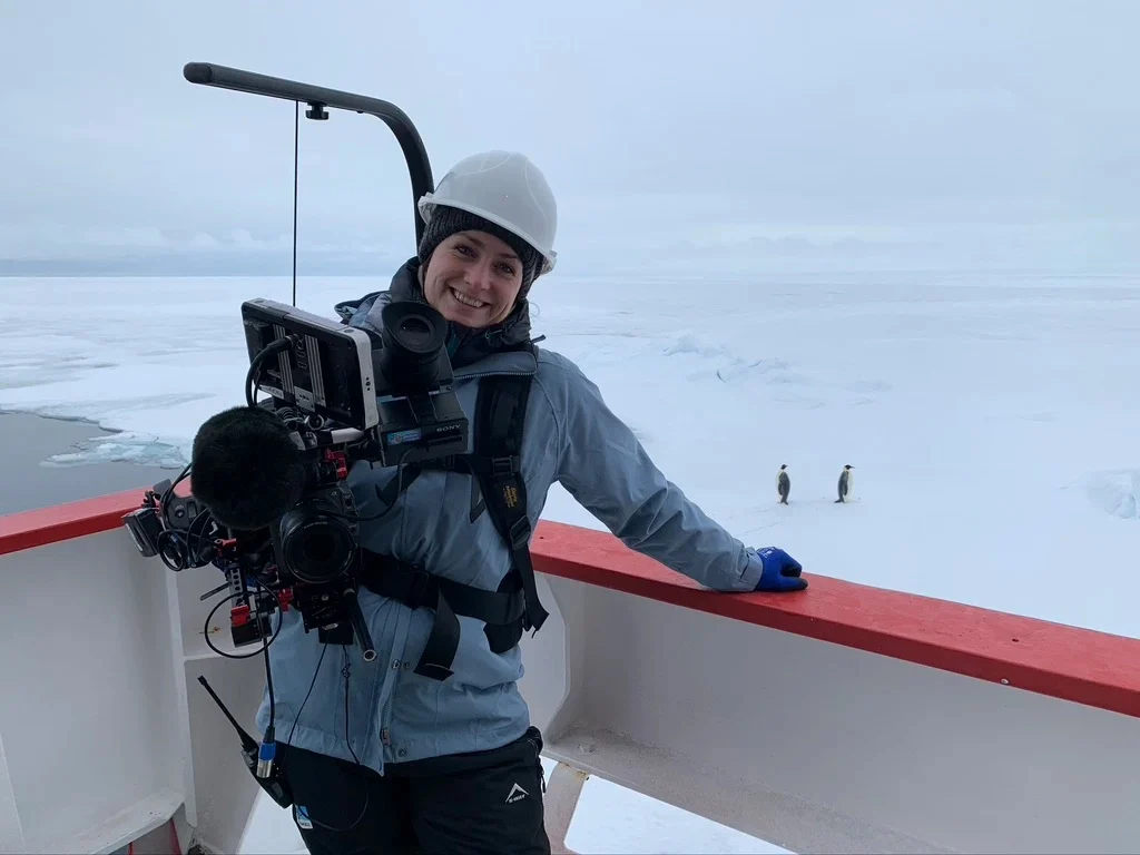 Natalie stood on the deck of a ship outside, with two penguins and Antarctica behind her. 