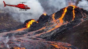 A volacno erupts with bright red and orange magma flowing down it's sides and a helicopter flies by to survey the scene.