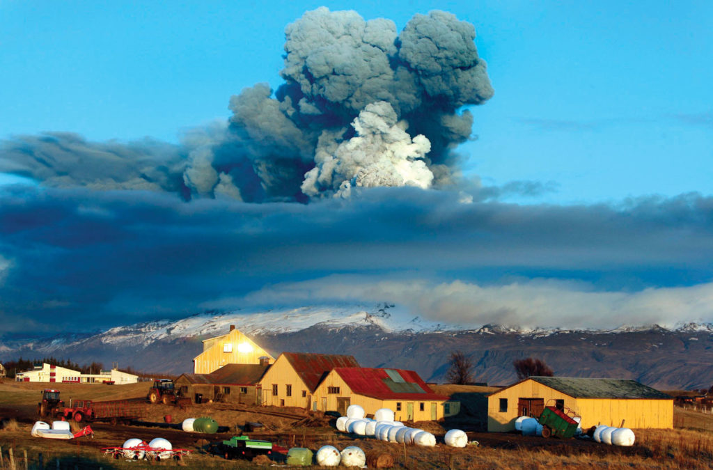 In the foreground there is a farm with a backdrop of a volcano is emitting large amounts of ash into the air.