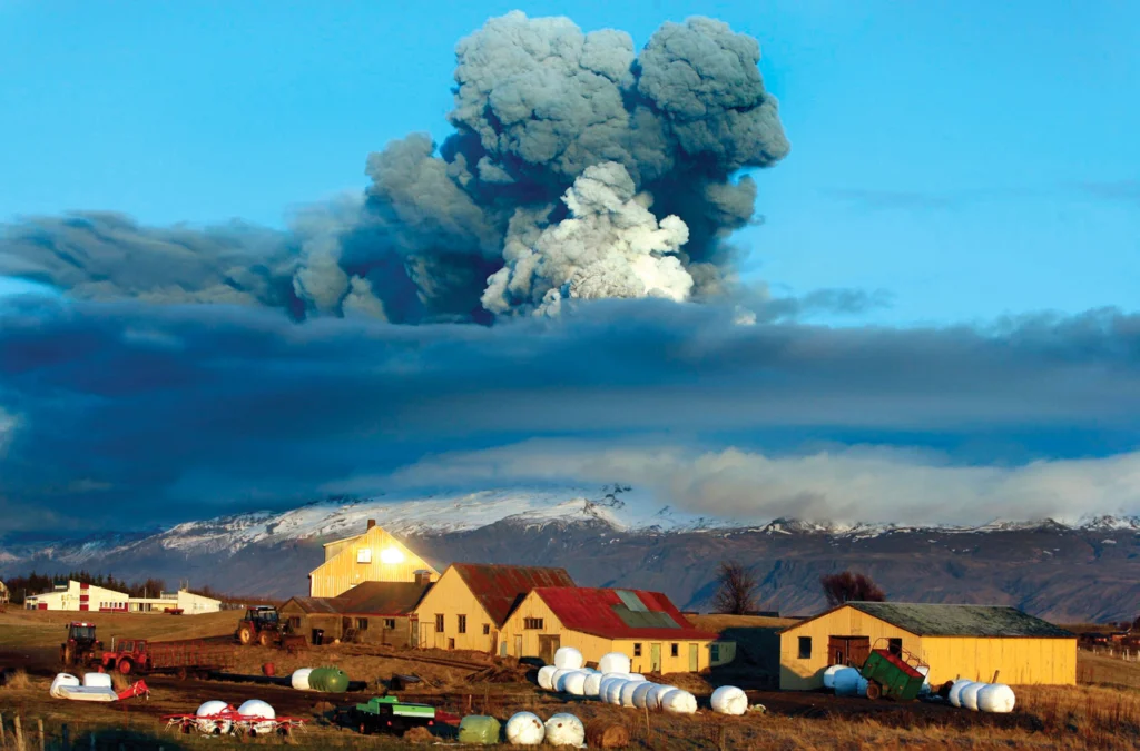 In the foreground there is a farm with a backdrop of a volcano is emitting large amounts of ash into the air.