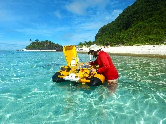 A photo of a tropical looking ocean, beach in background, clear water and blue skies. In the center of the image is a man wearing a red UV shirt. He is working on a large orange autonomous vehicle used for marine remote sensing.