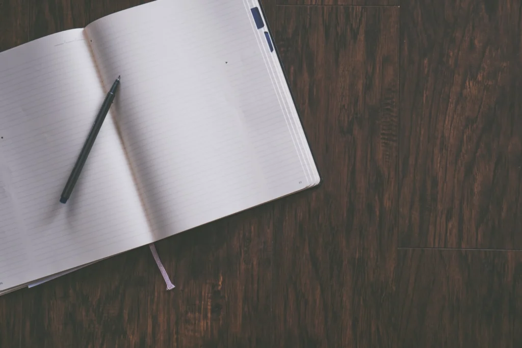 Dark table seen above with an open notebook on top of it. The notebook is empty and a pen lies on top of it.