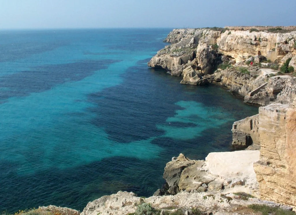 an image of a sea cove with rugged rocks to the right and bottom right of the image and clear blue sea water to the left and upper left of the image