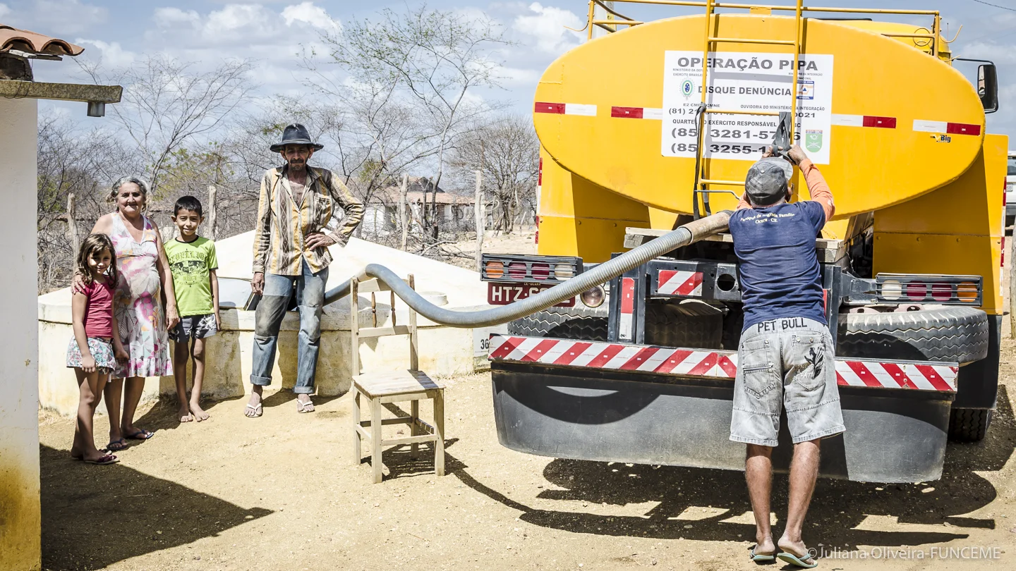 A family composed of mother, father, a young boy and a young girl on the left, a yellow water tank operated by a man filling the family's water tank.
